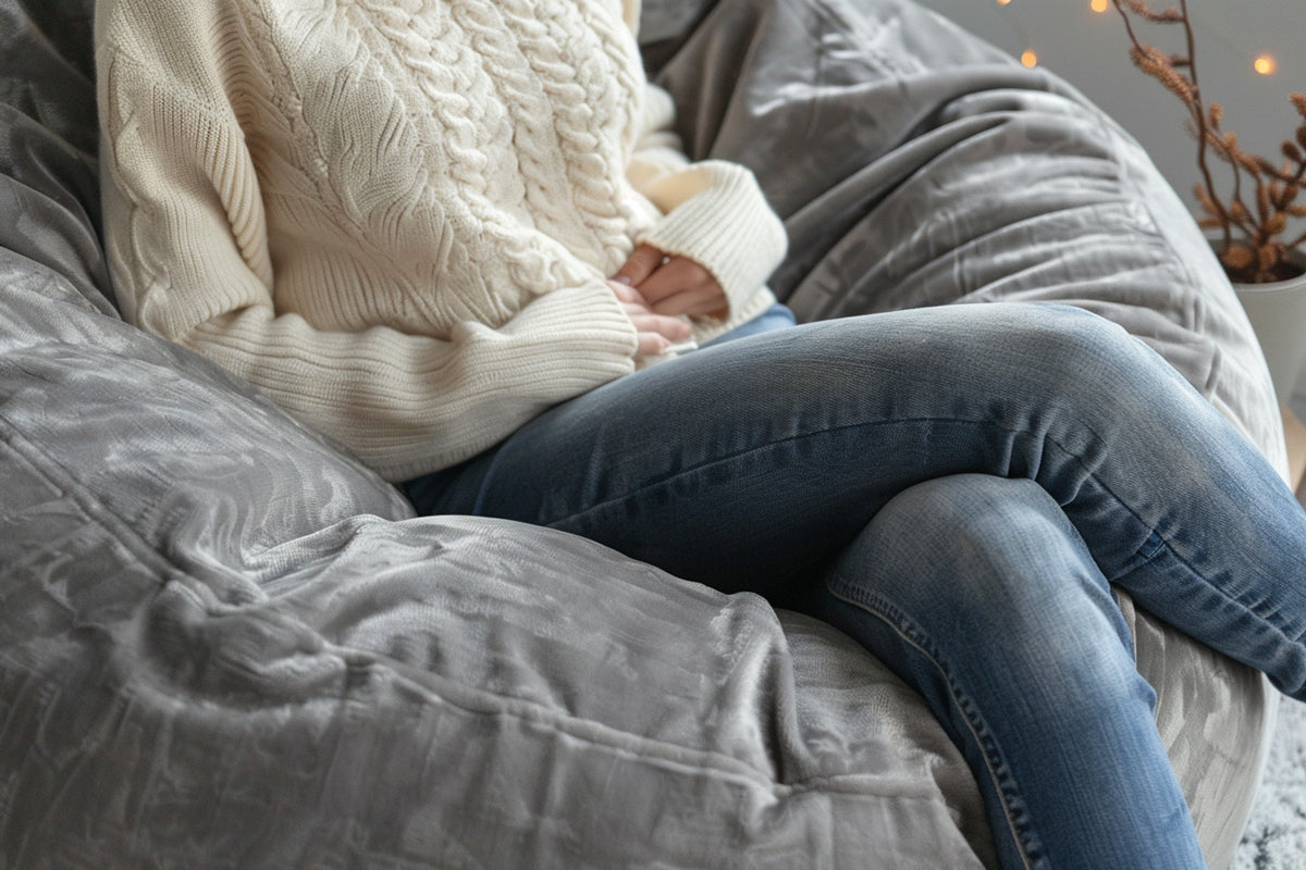 A woman is curled up and relaxing on a gray microsuede bean bag chair. She is wearing a cozy ivory sweater and blue jeans.