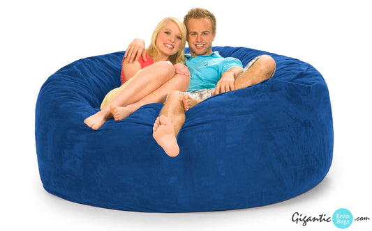 This shows our 6 ft round bean bag chair in blue. There is a happy couple resting on it wearing bright summer clothes. There is a logo on the bottom right displaying "Gigantic Bean Bags"