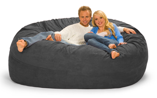 A 7 ft Charcoal Gray microsuede bean bag with a happy, cozy-looking couple in sweaters are relaxing on it.
