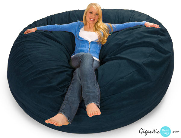 A blonde woman in a cozy sweater and jeans happily sitting on a 7 ft giant bean bag chair in Navy Blue.