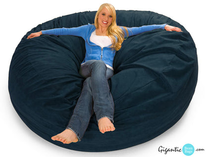 A blonde woman in a cozy sweater and jeans happily sitting on a 7 ft giant bean bag chair in Navy Blue.