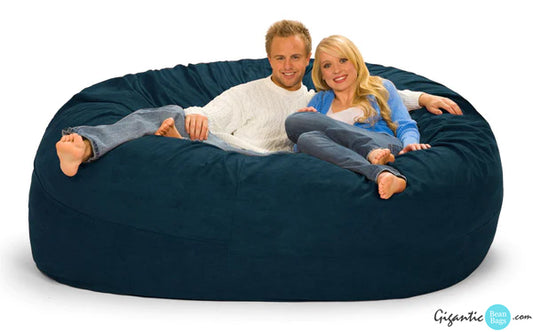 A 7 ft round navy blue bean bag chair with a couple wearing sweaters and jeans relaxing on it. 