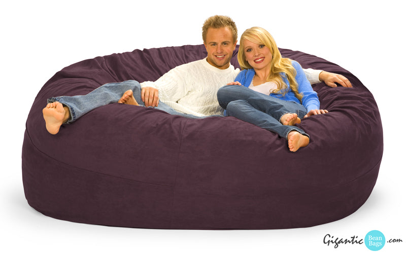 7 ft. bean bag with purple microsuede cover. Blonde couple chilling on it smiling at camera. White background. Logo at the bottom right.