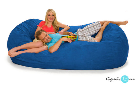 This image shows a royal blue oval bean bag lounger with a happy, relaxed, festive-looking couple on it. They are smiling at the camera and holding a bowl of chips. There is a white background and the Gigantic Bean Bags logo on the bottom right.