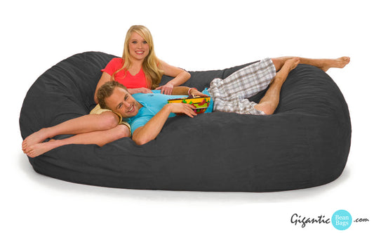 Our 7.5 ft gray bean bag lounger. This bean bag is an oval shape, has two people sitting on it, and displays the company logo on the bottom right.